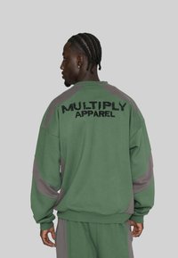 Mann mit geflochtenem Haar, der einen grünen und grauen Trainingsanzug mit dem Text "MULTIPLY APPAREL" auf dem Rücken trägt, seitlich vor einem grauen Hintergrund stehend.