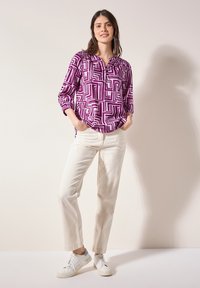 Blusa viola a maniche lunghe con un motivo geometrico, abbinata a pantaloni dritti color crema e sneakers bianche. Tessuto dalla texture liscia.