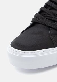 Černé tenisky s hladkým syntetickým svrškem, zaoblenou špičkou, silnou bílou podrážkou a zaoblenými tkaničkami. Obsahují jemné detailní prošívání.