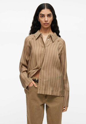 Vrouw in een losse, lichtbruine, verticaal gestreepte, knoopblouse met ruime manchetten en bijpassende bruine broek, staand met de hand in de zak.
