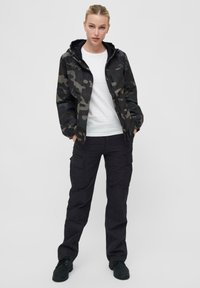 Camo pršiplášť v čiernych a zelených odtieňoch, so zipsom vpredu, kapucňou a elastickými manžetami; skombinovaný s čiernymi cargo nohavicami a tmavými topánkami.