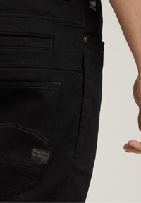 Pantalones de mezclilla negra con un acabado texturizado, que cuentan con múltiples bolsillos, un diseño cosido y una etiqueta de logo de marca en el costado.