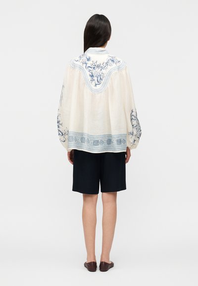 Γυναίκα που φοράει λευκό μπλουζάκι με μπλε κεντήματα με λουλούδια, μαύρες βερμούδες μέχρι το γόνατο και καφέ φλατ παπούτσια, στέκεται γυρισμένη με την πλάτη σε λευκό φόντο.