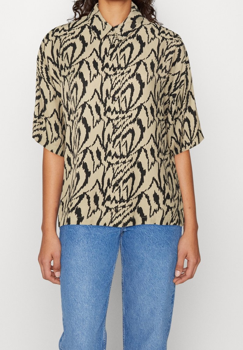 Personne portant une chemise courte beige et noire à motif abstrait, avec un col et un jean bleu.