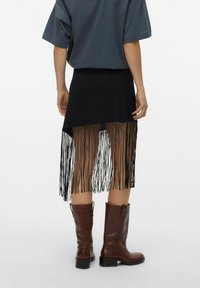 Jupe noire avec un ourlet à franges, associée à des bottes en cuir marron hautes jusqu'au genou. Le matériau semble doux, avec une chemise à coupe décontractée.