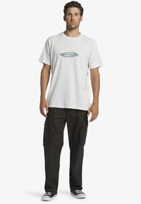 White cotton T-shirt with a blue logo on the chest, paired with dark green cargo pants featuring side pockets and flat-soled sneakers.