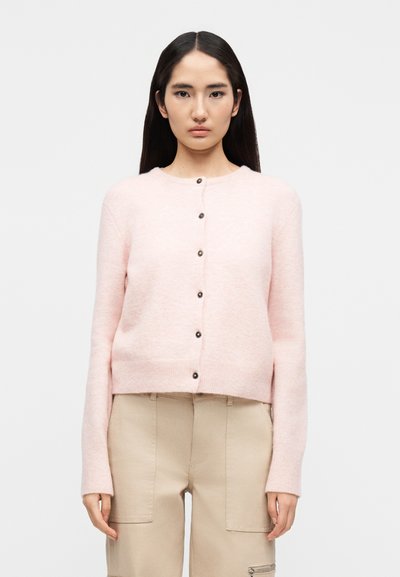 Femme debout devant un fond blanc, portant un cardigan rose clair boutonné et un pantalon beige avec des poches plaquées.