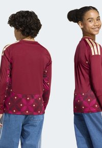 Red athletic long-sleeve shirt featuring gold stripes on shoulders and a geometric pattern in shades of pink and cream across the back.