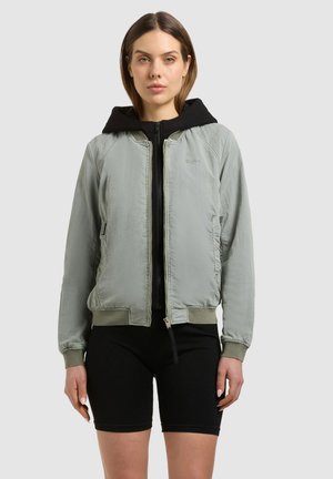 Frau trägt eine hellgraue Bomberjacke mit Reißverschluss über einem schwarzen Kapuzenpullover und schwarzen Radlerhosen, steht vor einem einfarbigen Hintergrund.