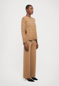 Maglione a coste beige con fessure laterali, abbinato a pantaloni beige a gamba larga. La modella indossa tacchi a punta neri, in piedi su uno sfondo bianco.