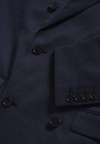 Mornarsko plavi blejzer od teksturirane tkanine, s crnim gumbima i detaljnim šavovima na rukavu. Uključuje džep na prsima.