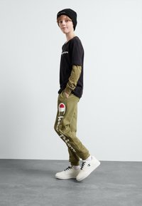T-shirt noir à manches longues superposé sur une chemise vert olive, associé à un jogging olive avec un logo, et des baskets blanches.