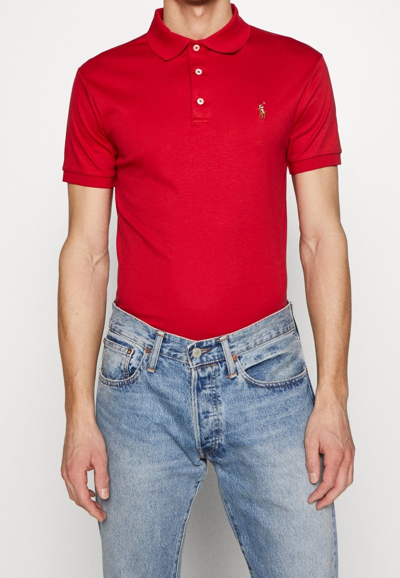 Homme portant un polo rouge vif à manches courtes rentré dans un jean délavé bleu clair, avec une main détendue le long du corps.