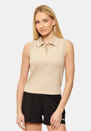 Mujer rubia con una camisa tipo polo beige sin mangas y pantalones cortos negros, de pie frente a un fondo liso.