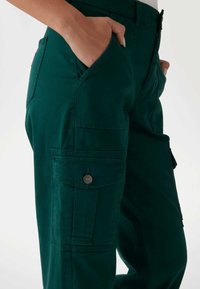 Zöld cargo nadrág, testhez álló fazonnal, oldalsó zsebekkel, elülső gombos bezáródással és puha, texturált anyaggal.