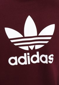 Vinröd sweatshirt med en vit treklöverlogotyp med tre blad ovanför ordet "adidas" i fetstil, gemener. Mjuk tygstruktur.