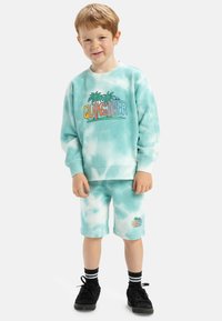Světle tyrkysová mikina s efektom tie-dye a logem "Quiksilver" spolu s palmami. Odpovídající šortky, oboje z měkkého materiálu. Černé boty s bílými pruhovanými ponožkami.