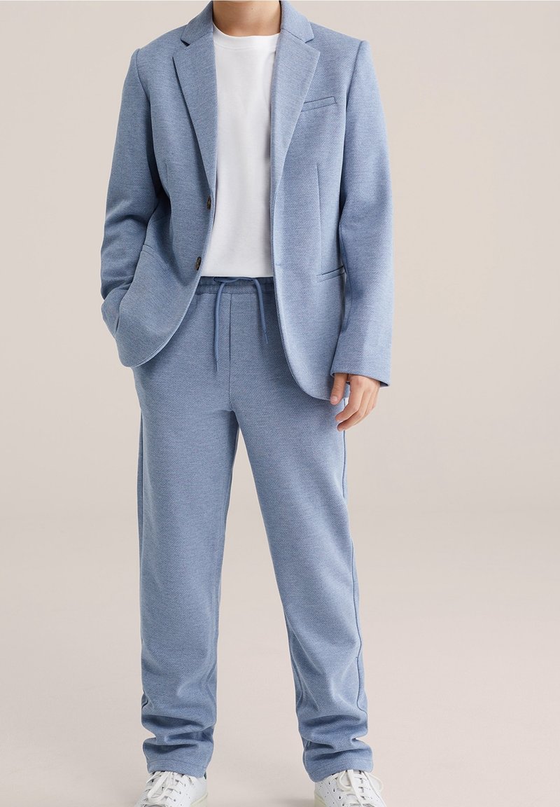 Personne portant une veste de costume bleu clair et un pantalon assorti avec un T-shirt blanc et des baskets blanches, debout devant un fond neutre.