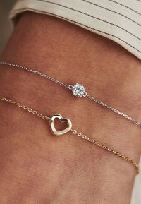 Silver and gold bracelets on a wrist; one features a round stone, the other has a heart shape, both with delicate chains.