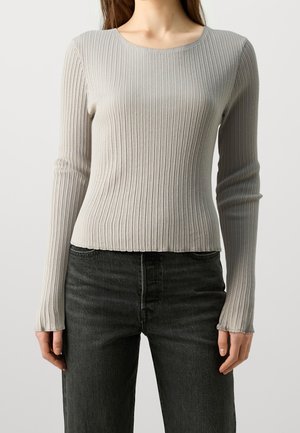 Femme portant un t-shirt à manches longues côtelé gris clair et un jean noir taille haute, debout devant un fond clair uni.