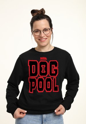 Kvinde med briller og hår i en knold, smilende, iført en sort sweatshirt med rød "DOG POOL" tekst og en knoglegrafik foran.