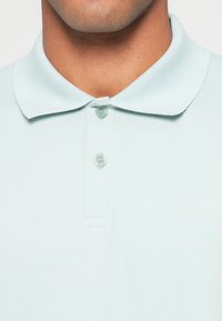 Világoskék galléros póló klasszikus gallérral, két gombbal és diszkrét logóval. Simább anyagtextúra és minimalista dizájn.