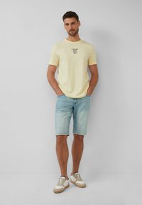 Hellgelbes T-Shirt mit kurzen Ärmeln und schwarzem Text, kombiniert mit hellblauen Jeansshorts. Das Model trägt beigefarbene und weiße Sneaker.