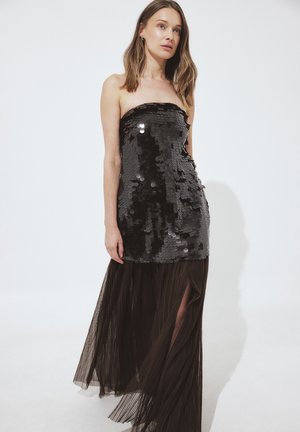 Zwart strapless jurk met een lijfje van pailletten en een vloeiende, doorschijnende zwarte geplooide rok met zijsplitten voor extra beweging en textuur.