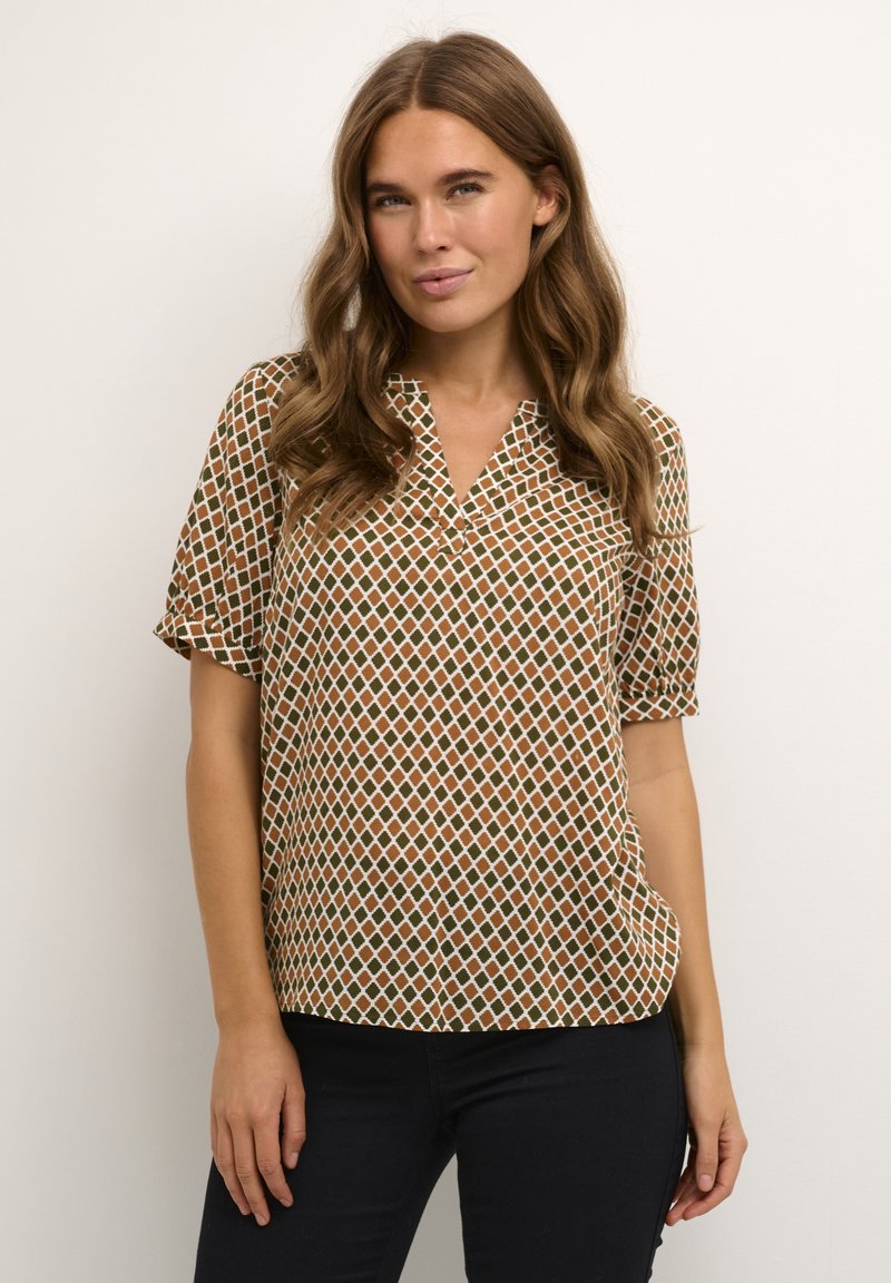 Femme aux longs cheveux bruns ondulés portant un chemisier marron et vert à manches courtes avec un motif en losanges et un pantalon noir, debout contre un mur beige.