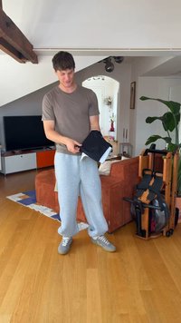 Νέος άνδρας με casual ρούχα διπλώνει ένα μαύρο ρούχο σε ένα μοντέρνο σαλόνι με μηχάνημα γυμναστικής και πορτοκαλί καναπέ.