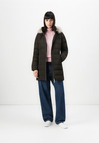 Ung kvinde iført sort pufferjakke med pelskrave, lyserød turtleneck, brede blå jeans og hvide sneakers, stående mod en hvid baggrund.