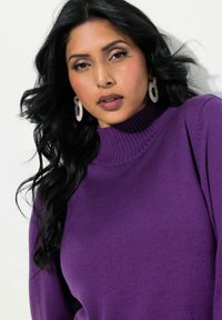 Maglione a collo alto viola con collo a costine, texture liscia e maniche aderenti. Orecchini di dichiarazione creano contrasto. Modella posa con lunghi capelli neri.