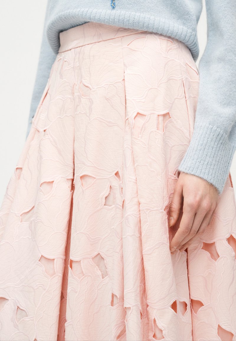 Gonna con motivo floreale rosa chiaro e dettagli ritagliati, abbinata a un maglione a maniche lunghe azzurro, con una mano parzialmente visibile di lato.