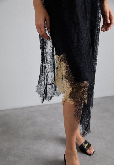 Μαύρο φόρεμα από δαντέλα με χρυσό τελείωμα από δαντέλα, που διαθέτει περίπλοκα σχέδια. Χαλαρός, ρο流η σχεδιασμός με επιμερισμένη υφή, συνδυασμένο με μαύρη υπόδηση.