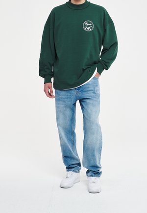 Persona vestida con un sudadera verde oscuro con un logotipo de montaña, jeans claros con un parche y zapatillas blancas, de pie contra un fondo sencillo.