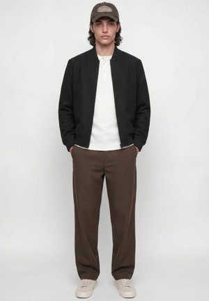 Jeune homme portant une veste noire zippée, une chemise blanche, un pantalon marron, des baskets beiges et une casquette marron, debout les mains dans les poches.