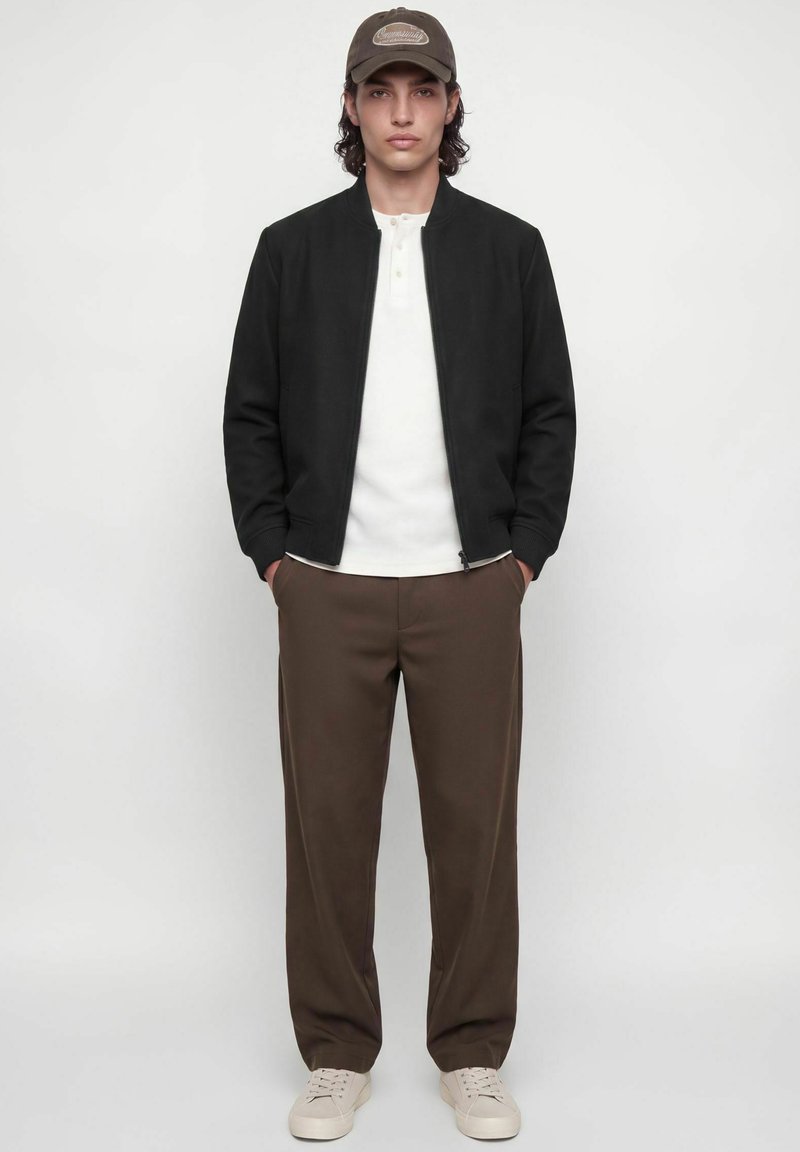 Jeune homme portant une veste noire zippée, une chemise blanche, un pantalon marron, des baskets beiges et une casquette marron, debout les mains dans les poches.