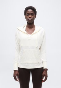 Γυναίκα που φοράει λευκό φούτερ με κουκούλα και τσέπη μπροστά, καθώς και σκουρόχρωμο κολάν, στέκεται μπροστά σε απλό φωτεινό φόντο.