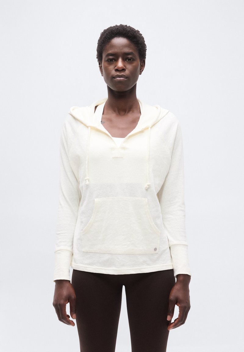 Γυναίκα που φοράει λευκό φούτερ με κουκούλα και τσέπη μπροστά, καθώς και σκουρόχρωμο κολάν, στέκεται μπροστά σε απλό φωτεινό φόντο.