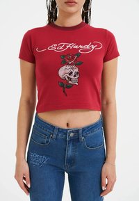 Rød bomulds crop-top med hvid "Ed Hardy" tekst og et design af et kranium med en rose. Kombineret med blå denim jeans med høj talje.