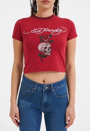 Red cotton cropped t-shirt featuring white "Ed Hardy" text and a skull with a rose design. Paired with blue denim high-waisted jeans.