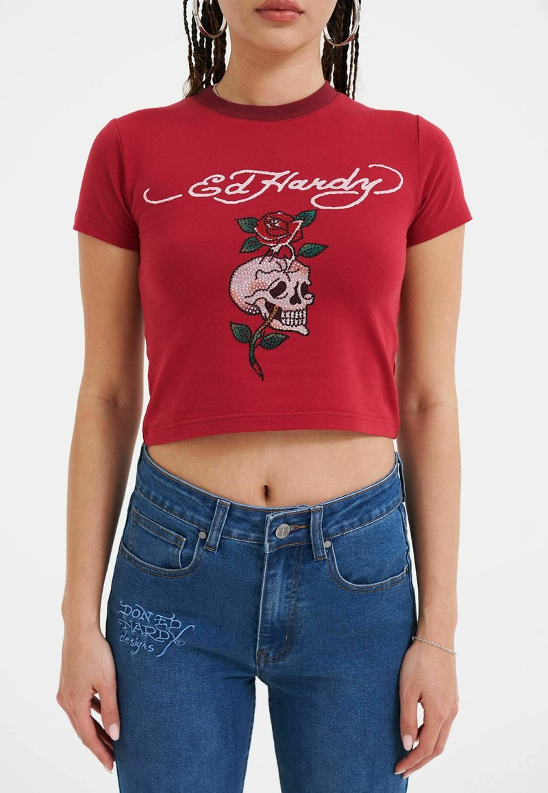 Rød bomulds crop-top med hvid "Ed Hardy" tekst og et design af et kranium med en rose. Kombineret med blå denim jeans med høj talje.