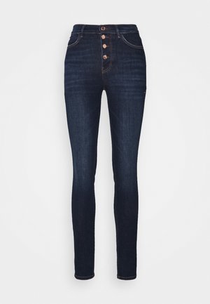 Jean skinny taille haute bleu foncé avec quatre boutons métalliques visibles et poches avant sur un fond clair uni.