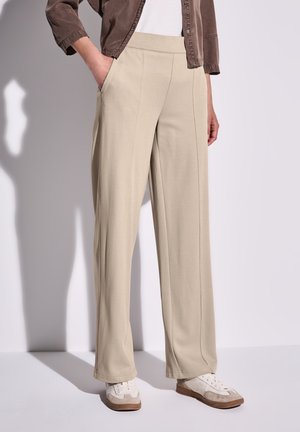 Person trägt beige weit geschnittene Hosen, weiße Sneakers und eine braune Jacke mit einer Hand in der Tasche vor einem einfarbigen Hintergrund.