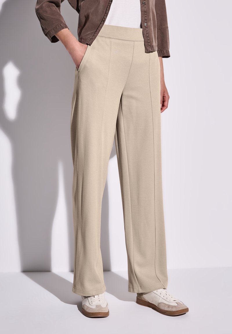 Personne portant un pantalon beige à jambes larges, des baskets blanches et une veste marron, une main dans la poche, sur un fond uni.