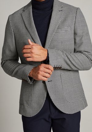 Mann richtet Manschette eines schwarz-weißen Hahnentrittmustersakko, das über einem schwarzen Rollkragenpullover und dunkler Hose getragen wird, vor neutralem Hintergrund.