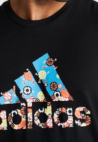 Schwarzes Baumwoll-T-Shirt mit einem leuchtend blauen Pixel-Art-Logo, das verschiedene sportbezogene Ikonen enthält, darunter Tennis und Basketball.