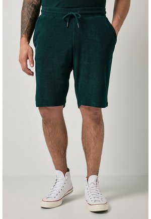 Uomo che indossa pantaloncini verde scuro con coulisse e sneakers bianche alte, una mano in tasca, tatuaggio visibile sul braccio sinistro.