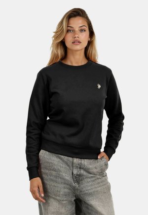 Woman with long blonde hair wearing a black sweatshirt with a small logo on the chest and light gray pants, one hand in pocket.