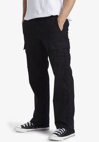 Black cargo pants made of cotton with a relaxed fit, featuring side pockets and a smooth texture. Worn with black sneakers and a white shirt.